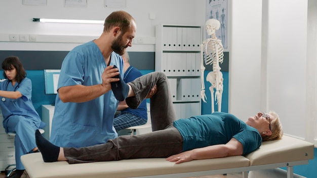 Male physician doing legs raise procedure with senior patient in orthopedic cabinet. Osteopath stretching muscles to increase mobility and cure elder woman with physical injury.