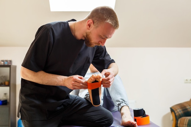 Male physiotherapist doctor massages the feet of a relaxed man sitting on a stretcher He uses kinesio tape Taping the foot