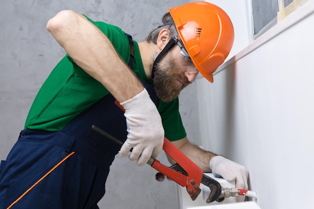 A male plumber installs a radiator in the heating system of an apartment Guy in overalls and a gas wrench