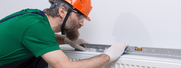 A male plumber installs a radiator in the heating system of an apartment Guy in overalls and a gas wrench