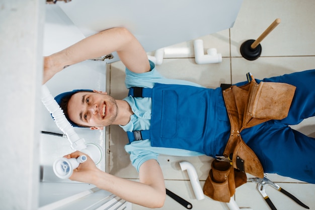 Male plumber in uniform installing drain pipe in the kitchen, top view. Handywoman with toolbag repair sink, sanitary equipment service at home