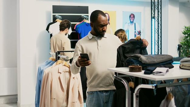 Male shopper looking at online website to find clothes, buying new fashionable merchandise in clothing store. Person with smartphone checking stock of formal or casual wear, shopping mall.