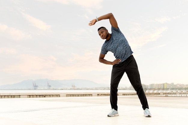 Male in sportswear standing near sea while looking away and doing stretching exercises