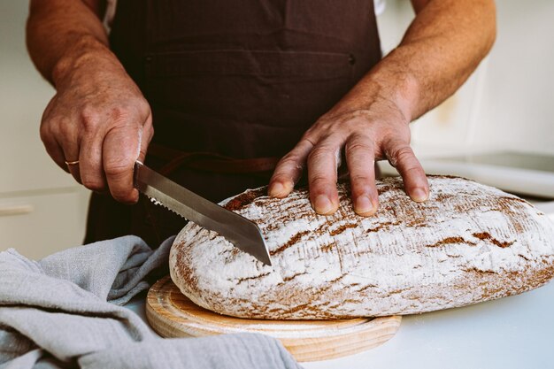 Male strong muscular hands of baker, in flour, cut loaf of craft homemade bread from rye flour