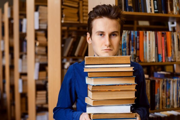 Photo male student stands with a pile of books at the library looks very disappointed