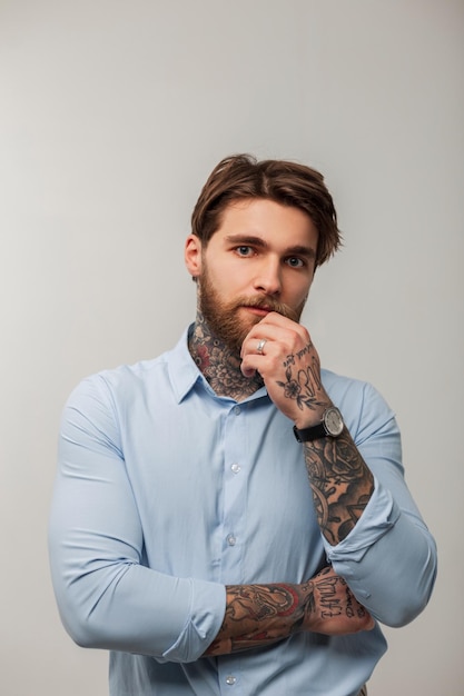 Male studio portrait of a handsome brutal man with a hairstyle and a beard with a tattoo in a blue fashion shirt on a gray background