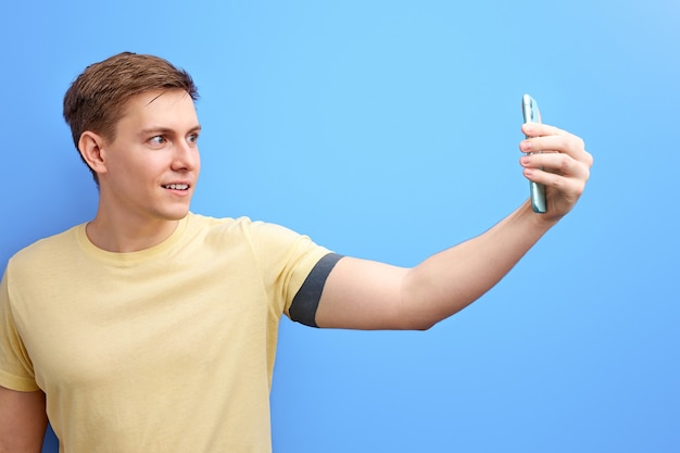 Photo male taking photo on smartphone, looking at phone's camera and smiling, isolated over blue background. studio portrait