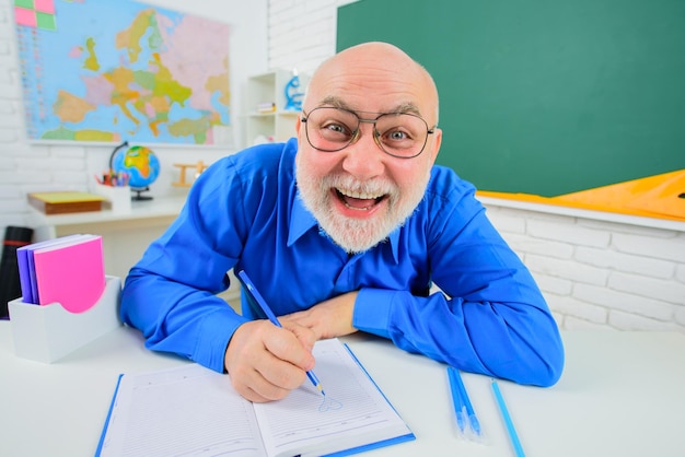 Male teacher sitting in school classroom teacher working at his desk happy tutor with notebook in