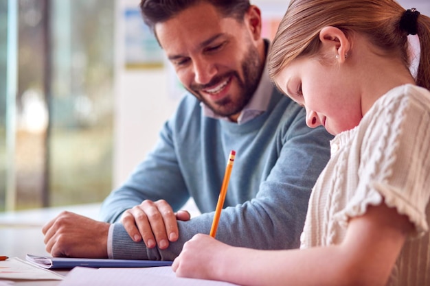 Male Teacher With Female Student In School Classroom Sitting At Desk Writing In Book Together