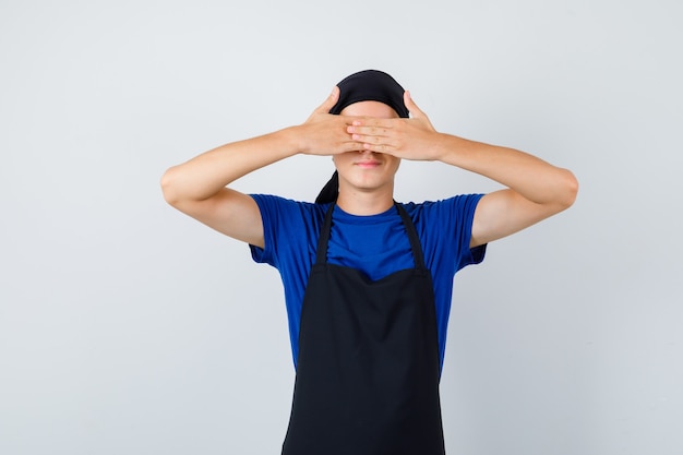 Male teen cook covering eyes with hands in t-shirt, apron and looking ashamed , front view.