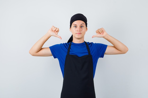 Male teen cook pointing at himself with thumbs in t-shirt, apron and looking confident. front view.