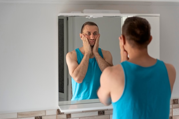 Male touching his beard and face Sleepy man in bathroom looks at himself in mirror Morning routine