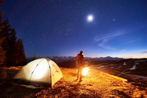 Male tourist have a rest in his camp at night, standing near campfire and tent under beautiful night sky full of stars and the moon and enjoying night scene in the mountains