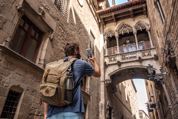 A male traveler with a backpack takes a selfie with a mask while visiting the Gothic quarter of the city of Barcelona, Spain