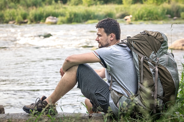 A male traveler with a large hiking backpack sits resting near the river.