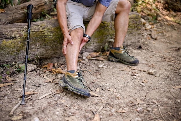 A male trekker sits on a wooden log and massages his leg suffering from knee pain leg cramping