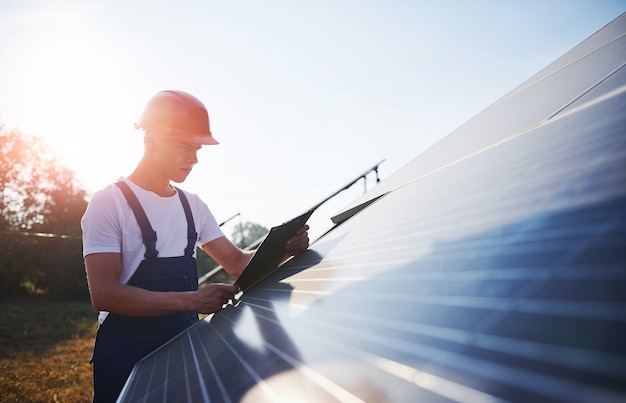 Male worker in blue uniform outdoors with solar batteries at sunny day.