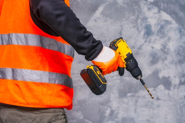 Male worker holds a closeup electric cordless screwdriver in his hands against the background of a construction tool and a concrete wall