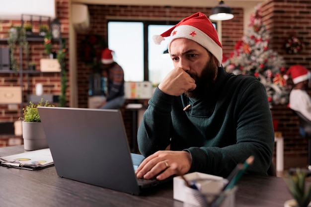 Male worker using laptop in business office to work on startup project during winter season. Man with santa hat working on report in workplace with christmas tree and holiday decorations.