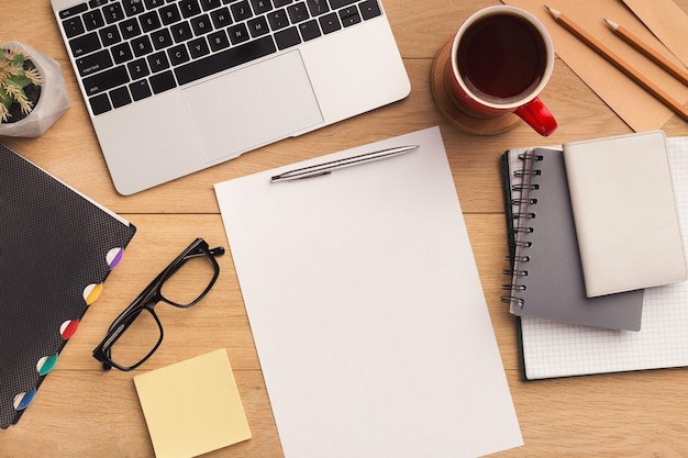 Male workplace with empty sheet of paper for advertisement, laptop, coffee cup, notebook and glasses. Wooden background, top view, mockup, copy space.