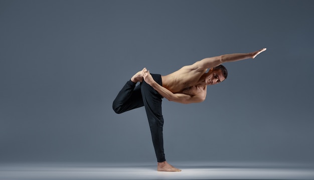 Male yoga keeps balanc in a difficult pose on one leg, grey background. Strong man doing yogi exercise, asana training, top concentration, healthy lifestyle