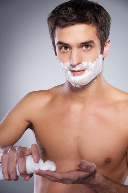 Man applying shaving cream. Handsome young shirtless man applying shaving cream at his face and looking at camera while standing isolated on grey background