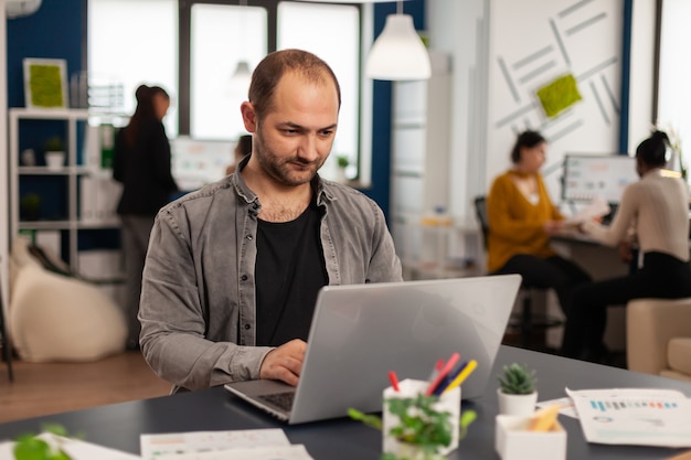 Man authentic manager sitting at table in modern startup business office sending mails typing on laptop