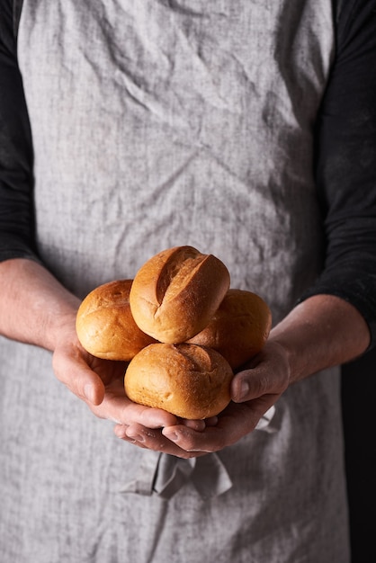 A man baker with a beard in a gray apron stands against a black background and holds, breaks, cuts off delicious, crispy bread, rolls, baguette.