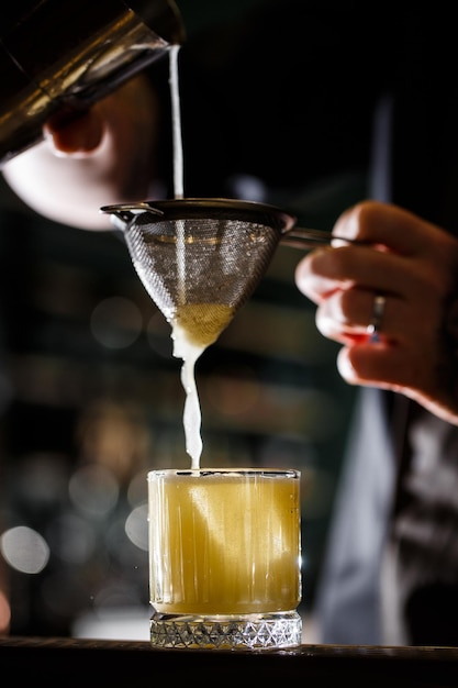 Man bartender carefully filters bright alcoholic cocktail from steel shaker cup into glass through sieve Closeup view