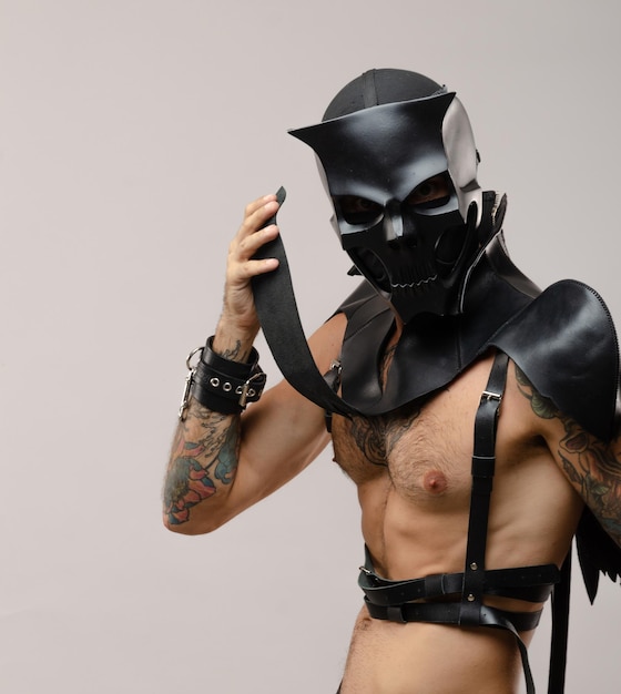 A man in a bdsm demon skull mask dressed in a leather cloak with leather bracelets and straps on his body