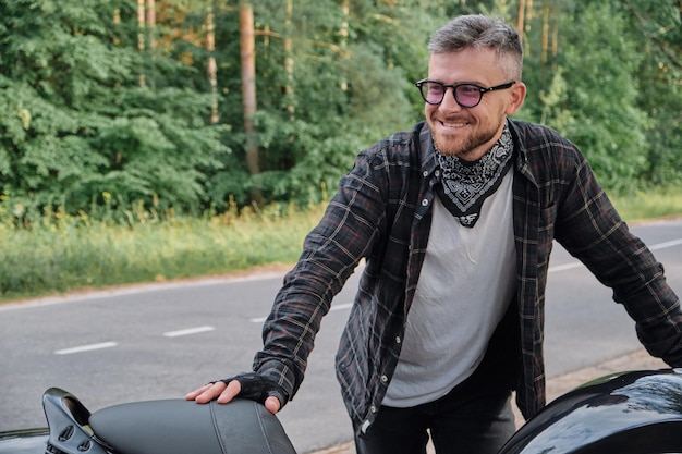 Man biker middle aged motorcyclist smiling stands with a motorcycle on the side of the road in the f