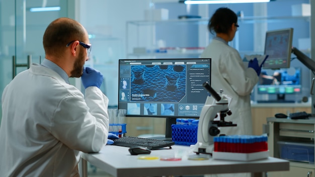 Man biologist checking DNA informations typing on pc in modern equipped laboratory. Scientists examining vaccine evolution in medical lab using high tech, chemistry tools for scientific research.