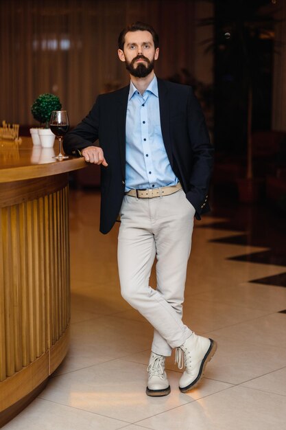 Photo a man in a black blazer stands in front of a wooden bar with a glass of wine.