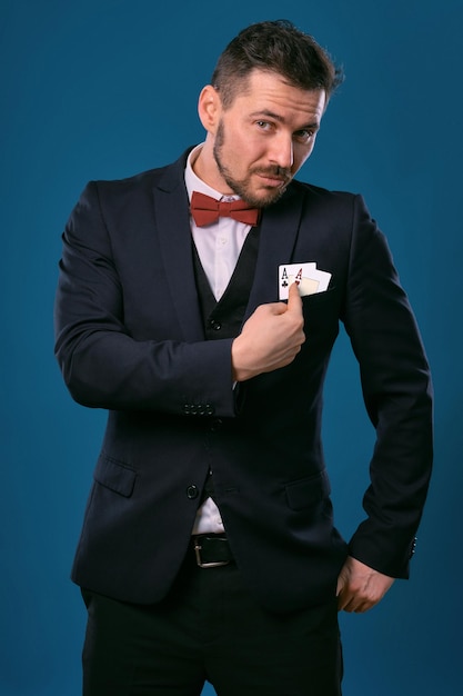 Man in black classic suit and red bowtie showing two playing cards while posing against blue studio ...