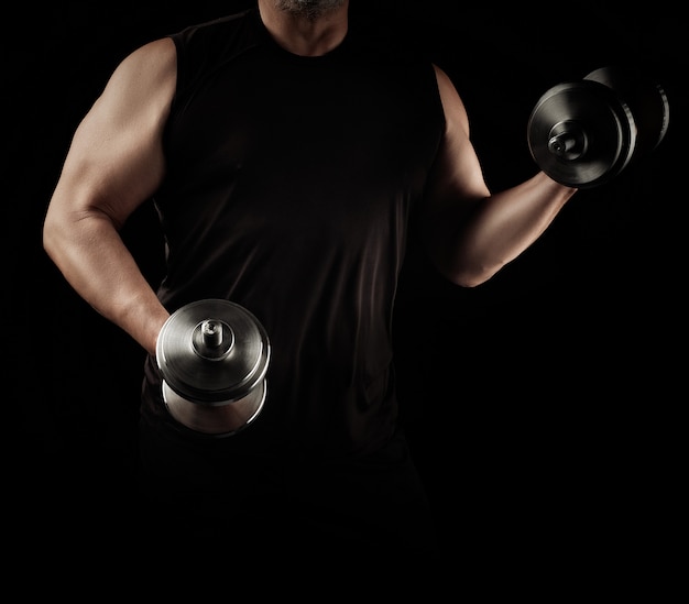 Man in black clothes holds steel dumbbells in his hands, his muscles are tense