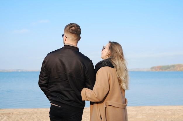 A man in a black jacket and a woman in a warm brown coat stand with their backs to the camera and look at the lake