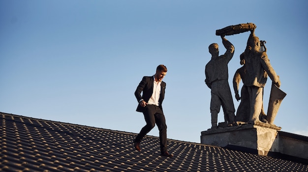 Photo man in black luxury clothes walk on the rooftop near the statue.