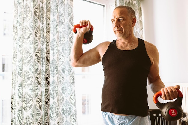 A man in a black shirt and blue pants lifts dumbbells in front of a window.