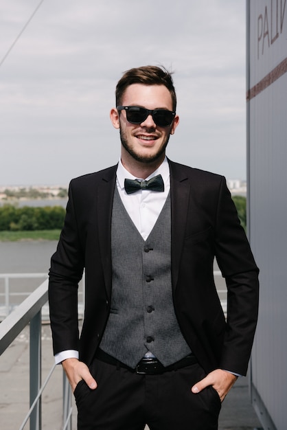 A man in a black suit and a black bowtie poses in the open air. Advertise menswear.