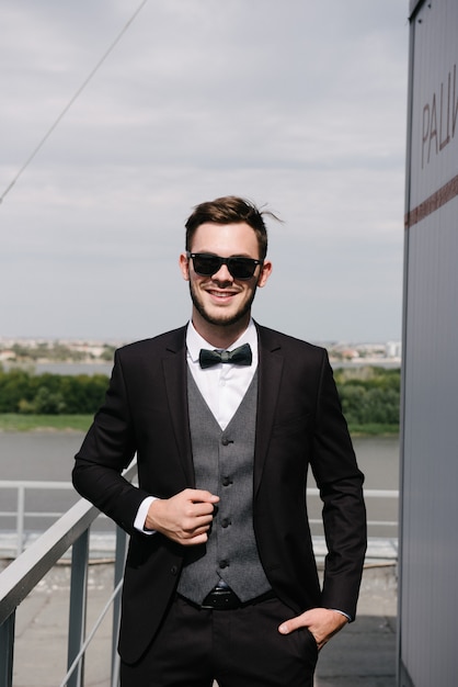 A man in a black suit and a black bowtie poses in the open air. Advertise menswear.