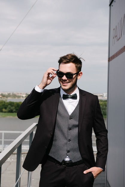 A man in a black suit and a black bowtie poses in the open air.