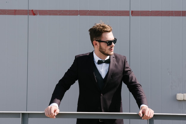 A man in a black suit and a black bowtie poses in the open air.