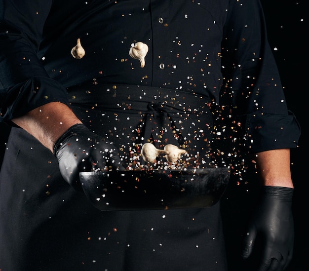 Photo man in a black uniform holding a round cast iron pan with salt and pepper chef tosses spice u