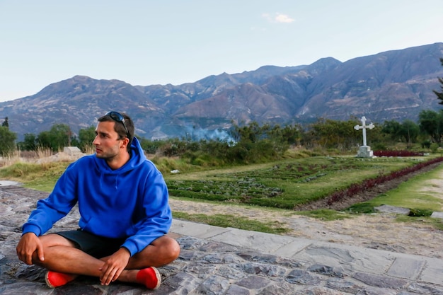 Man in a blue shirt sitting on stones in a green field and trees at sunset.