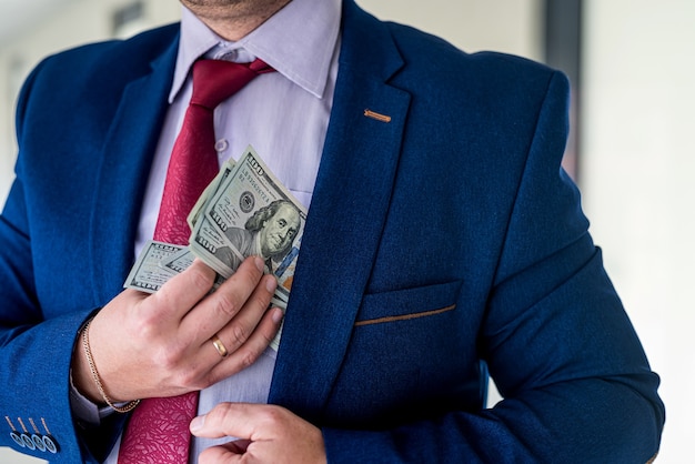 Man in blue suit and red tie putting bribe dollar bills into his pocket. corruption concept