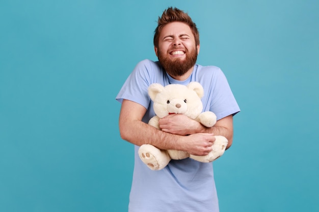 Man in blue Tshirt standing with closed eyes and toothy smile embracing white soft teddy bear