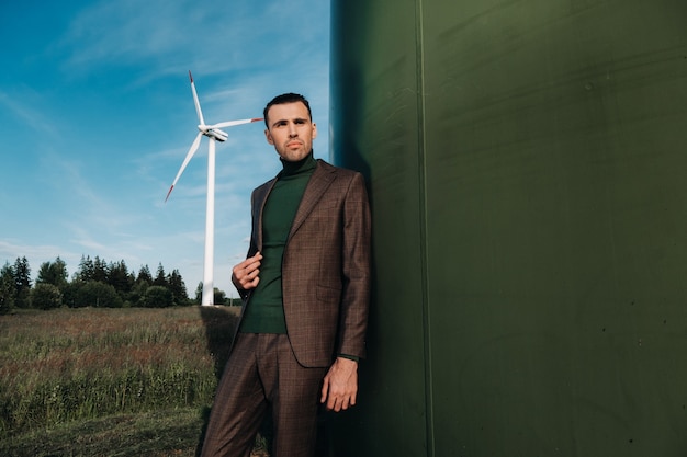 A man in a business suit with a green Golf shirt stands next to a windmill against the background of the field and the blue sky.Businessman near the windmills.Modern concept of the future.