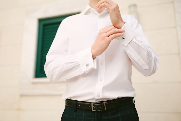 Man buttons up the cuffs of a white shirt against the backdrop of a wall with a window closeup