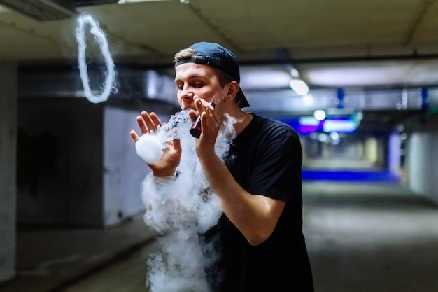 Man in cap smoke an electronic cigarette and releases clouds of vapor