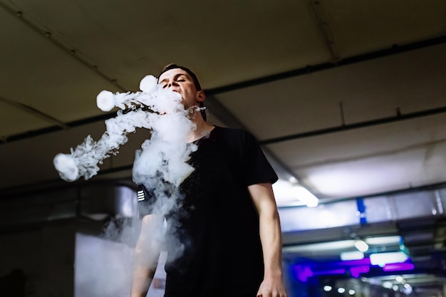 Man in cap smoke an electronic cigarette and releases clouds of vapor